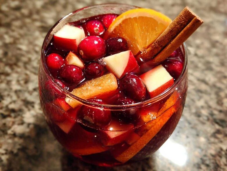 Close-up of a glass filled with rich red holiday sangria, loaded with cranberries, apple chunks, and an orange slice garnished with cinnamon sticks.
