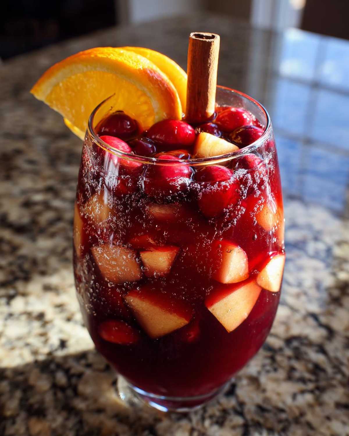Close-up of a glass filled with dark red holiday sangria, loaded with cranberries, apple chunks, and garnished with an orange slice and cinnamon stick.