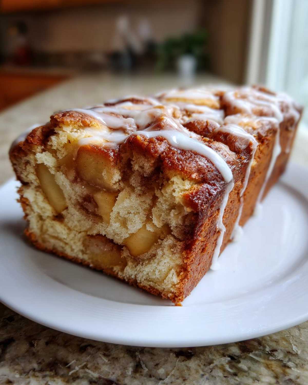 A close-up of a loaf of moist apple fritter bread, studded with apple chunks and topped with a white vanilla glaze.