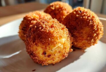 Close-up of four golden brown, crispy fried rice balls resting on a white plate in bright sunlight.