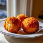 Three crispy, golden brown rice balls served on a white plate, with one showing the creamy rice interior.
