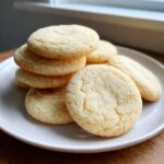 A stack and arrangement of freshly baked, pale yellow soft sugar cookies coated in granulated sugar.