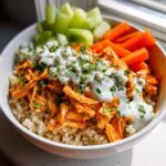 Close-up of a healthy buffalo chicken bowl featuring shredded chicken in orange sauce over brown rice, topped with blue cheese dressing and crumbles.
