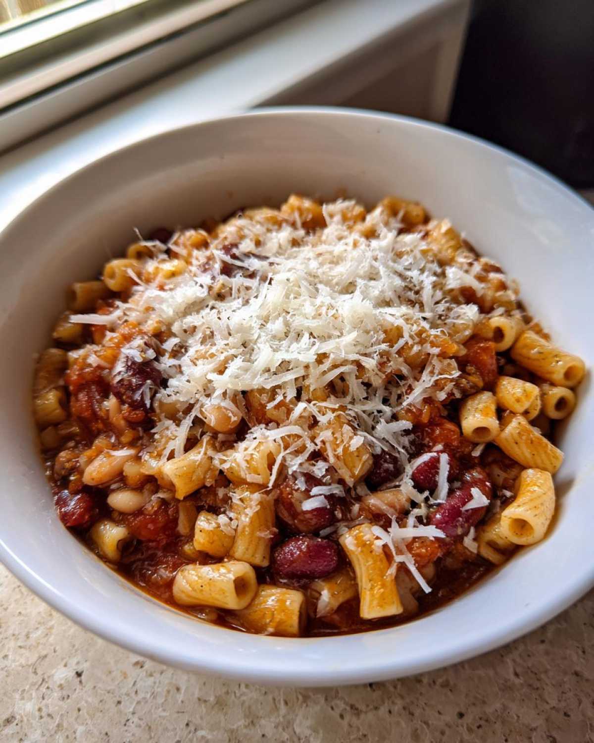 A close-up of a white bowl filled with rich pasta fagioli, topped generously with grated Parmesan cheese.