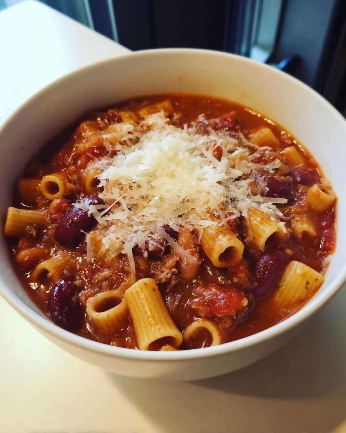 Close-up of a white bowl filled with rich pasta fagioli, featuring rigatoni pasta, beans, and topped with grated Parmesan cheese.
