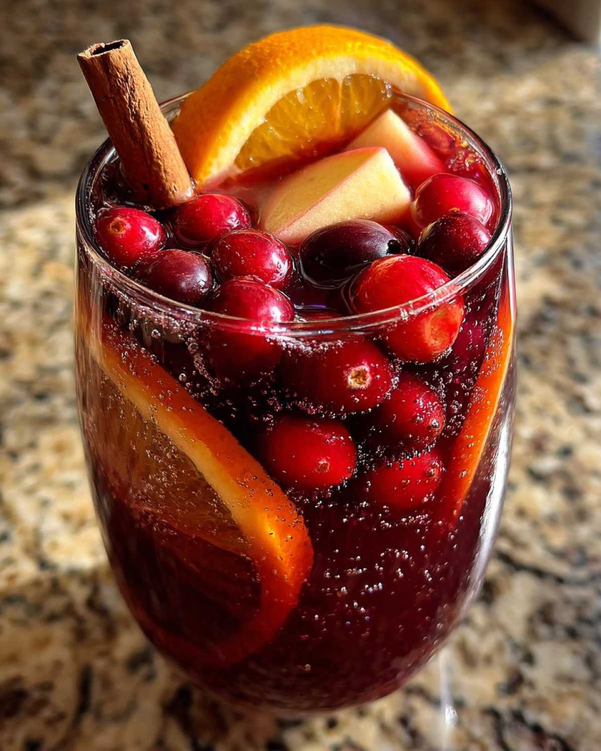 Close-up of a glass filled with bubbly, dark red holiday sangria garnished with fresh cranberries, orange slices, apple chunks, and a cinnamon stick.