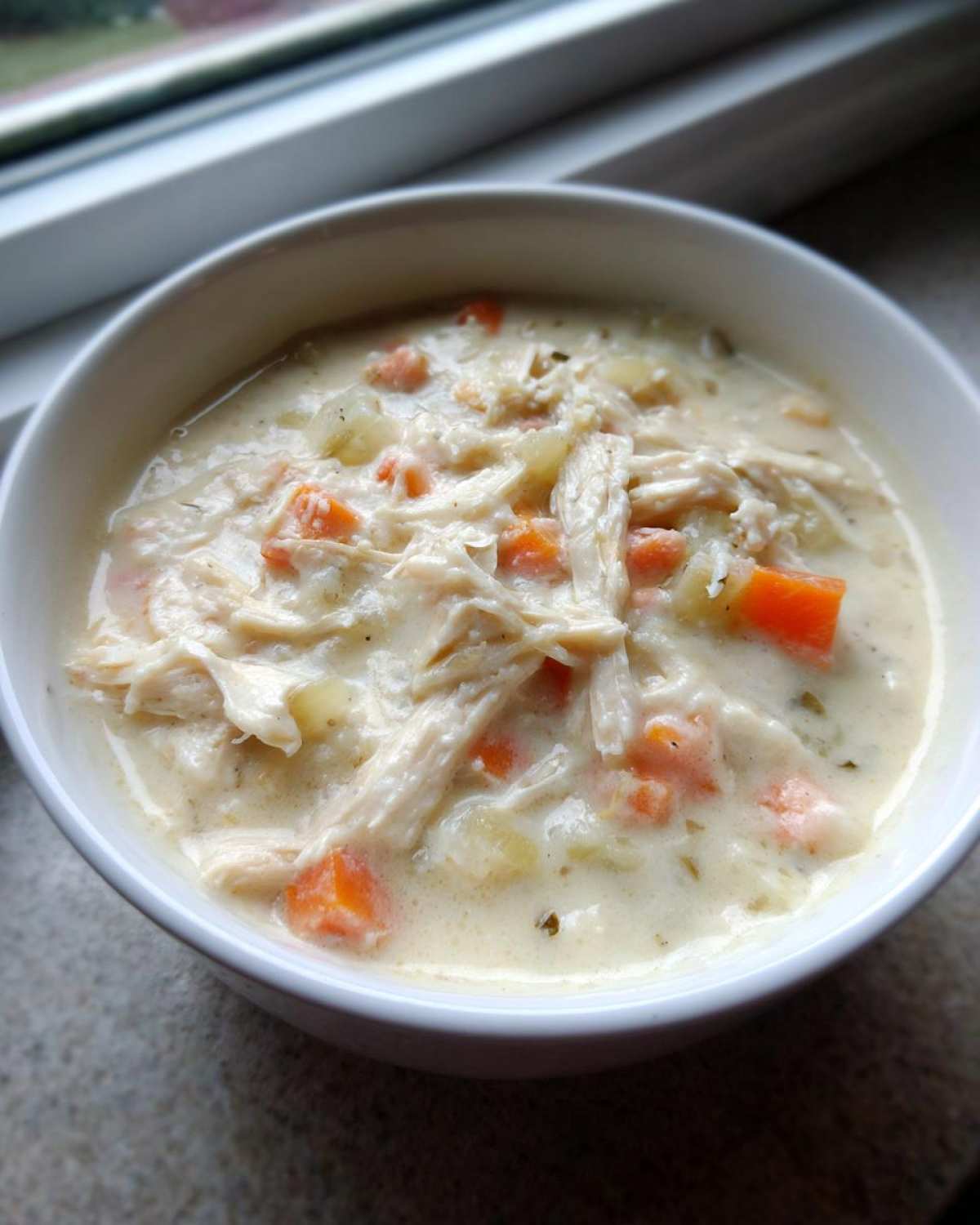 Close-up of a white bowl filled with hearty homemade cream of chicken soup featuring shredded chicken and diced carrots.