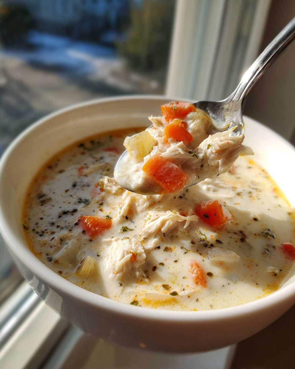 A spoonful lifted from a bowl of homemade cream of chicken soup, showing shredded chicken, carrots, and herbs.