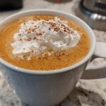 Close-up of a homemade pumpkin spice latte in a white mug, topped with whipped cream and cinnamon.
