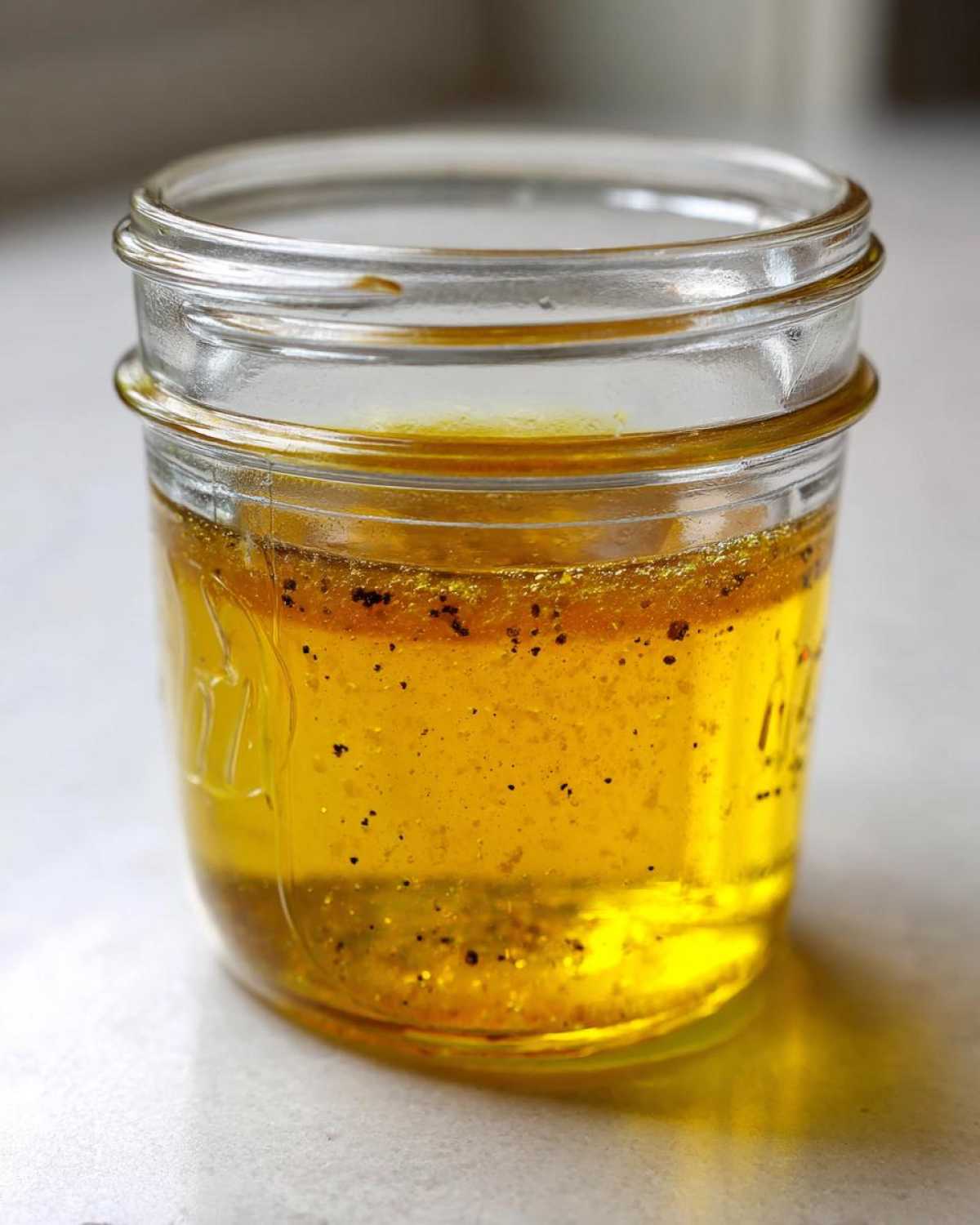 Close-up of separated homemade salad dressing, a yellow oil layer floating over spices in a small glass jar.