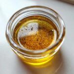 Close-up of homemade salad dressing, a golden vinaigrette with visible black pepper and spices, inside a small glass jar.