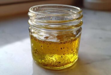 Close-up of homemade vinaigrette salad dressing, showing golden oil and dark spices settled in a small glass jar.