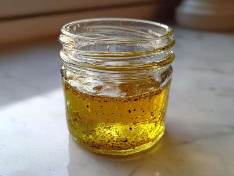 Close-up of homemade vinaigrette salad dressing, showing golden oil and dark spices settled in a small glass jar.