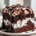 Close-up of a decadent slice of hot chocolate poke cake showing layers of dark chocolate cake, white filling, and chocolate whipped topping with fudge drizzle.