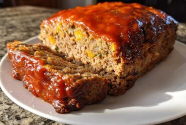 A freshly baked meatloaf with a thick, shiny glaze, showing one slice cut and revealing a moist interior.