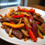 Close-up of juicy steak fajitas featuring sliced steak, colorful bell peppers (red, yellow, green), and caramelized onions on a white plate.