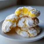 A stack of three delicious lemon crinkle cookies heavily dusted with powdered sugar, one cookie is broken open showing the bright yellow interior.