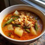 Close-up of a bowl of rich, reddish-orange Maryland crab soup filled with chunks of crab meat, potatoes, green beans, and corn.