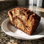 A moist, thick slice of apple bread showing chunks of apple and a cinnamon swirl, served on a white plate.