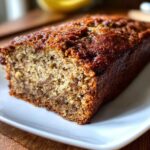 Close-up of a freshly baked, moist banana bread loaf with a dark, caramelized crust, resting on a white plate.