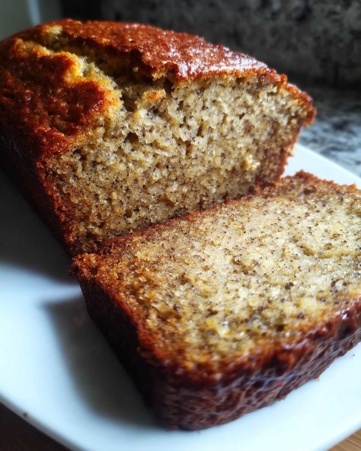 Close-up of a moist banana bread loaf, showing a thick slice cut and resting against the main loaf on a white plate.