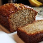 Close-up of a moist banana bread loaf with one slice cut and resting beside it on a white plate.
