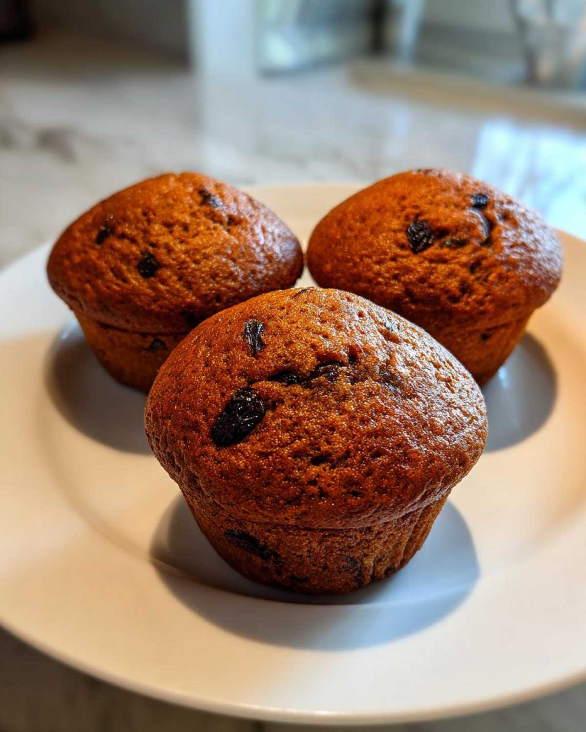 Three freshly baked, golden-brown bran muffins studded with raisins resting on a white plate.