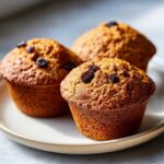 Close-up of three freshly baked, golden-brown bran muffins topped with dark raisins, sitting on a light ceramic plate.