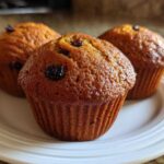 Close-up of three golden brown, moist bran muffins topped with raisins, resting on a white plate.