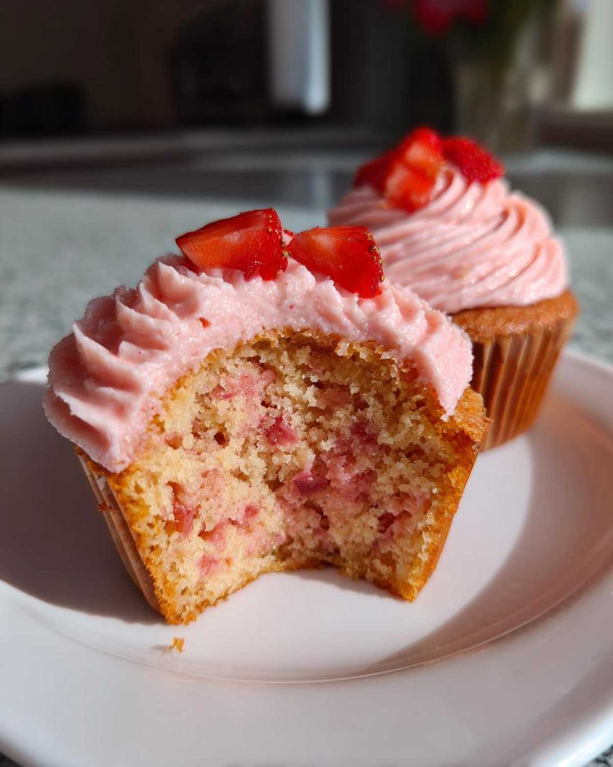 Close-up of a moist strawberry cupcake with a bite taken out, showing pink frosting and strawberry pieces inside.