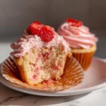 Close-up of a strawberry cupcake cut in half showing the moist interior and pink frosting, topped with fresh strawberry slices.