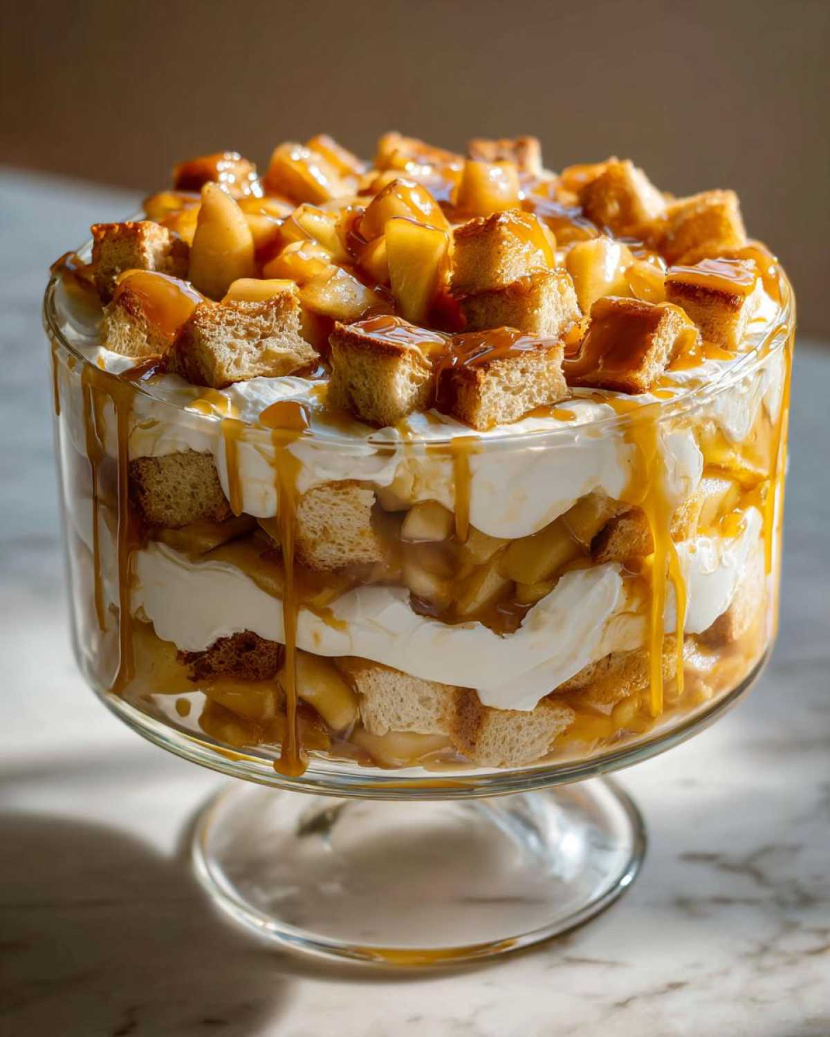 Layers of cake, cream, and spiced apples topped with cubes and drizzled with caramel in a glass caramel apple trifle.
