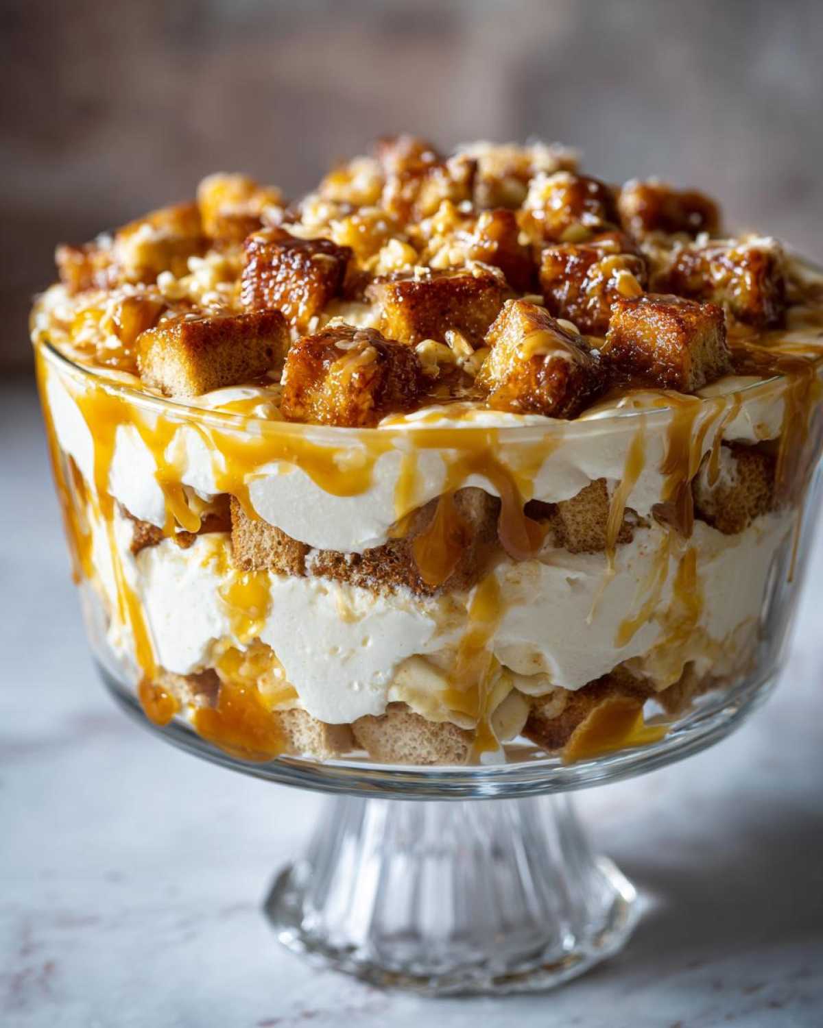 A beautiful, layered no-bake caramel apple trifle featuring cream, cake pieces, and caramel drizzle in a glass pedestal dish.
