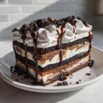 A tall, layered slice of no-bake ice cream cake featuring dark chocolate layers, creamy fillings, whipped topping, and chocolate drizzle.