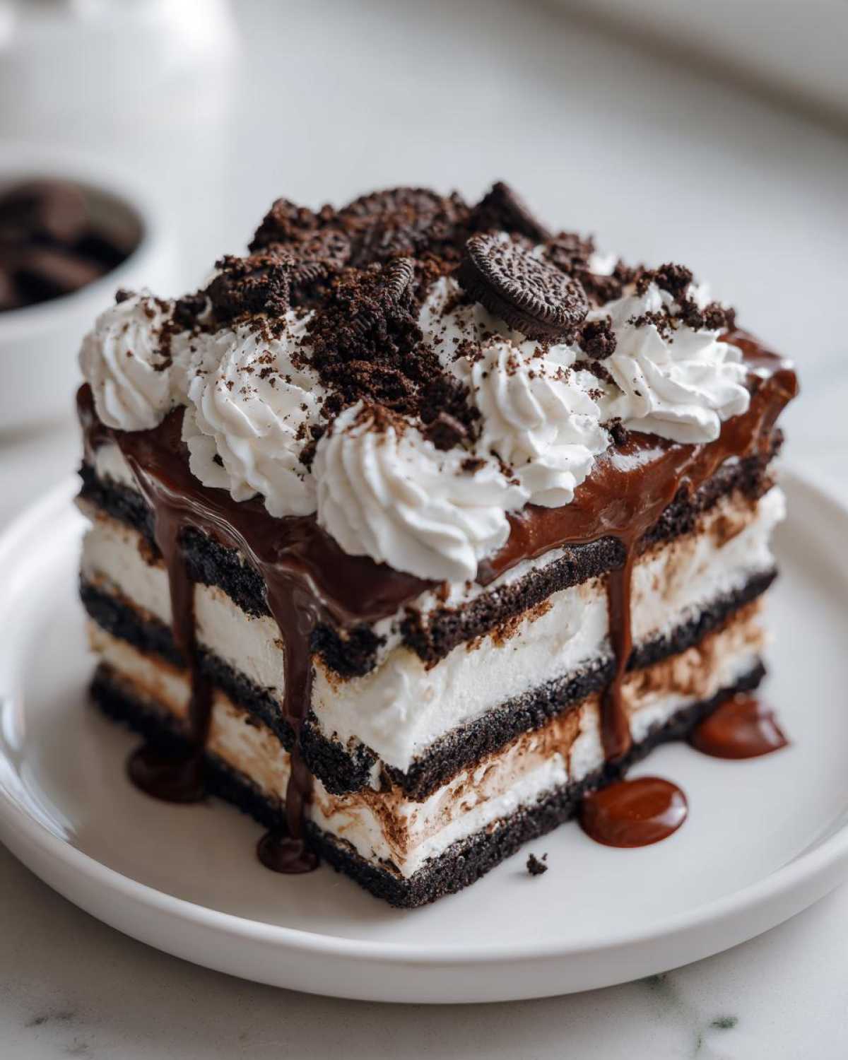 A decadent square slice of no-bake ice cream cake layered with chocolate cookies and cream filling, topped with whipped cream and fudge.
