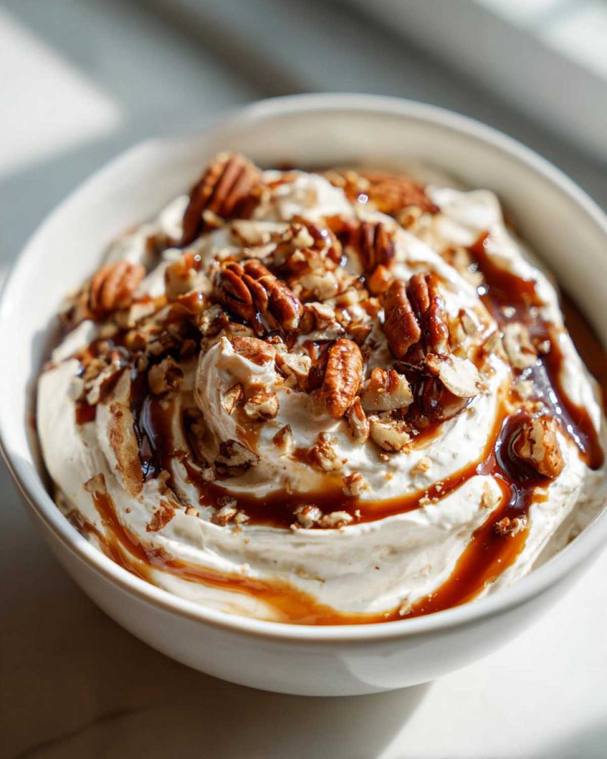 A close-up of creamy no-bake pecan pie dip topped with pecans and caramel sauce in a white bowl.