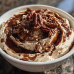 A close-up of a creamy no-bake pecan pie dip topped with whole pecans and caramel sauce.