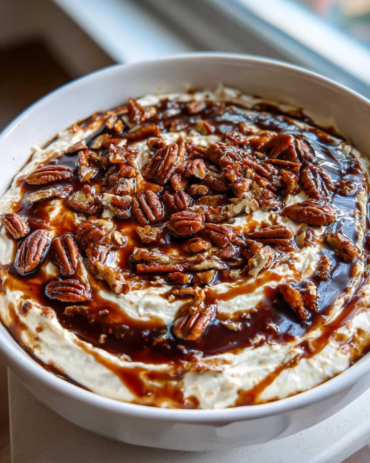A white bowl filled with creamy no-bake pecan pie dip, generously topped with caramel sauce and whole pecans.