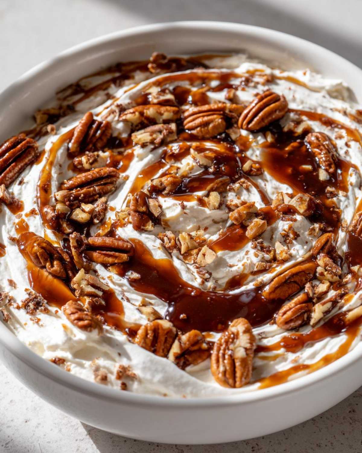 A bowl of creamy, no-bake pecan pie dip topped with whole pecans, chopped pecans, and a rich caramel drizzle.