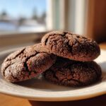 Three dark, crinkled ginger molasses cookies coated in sugar resting on a light-colored plate near a window.