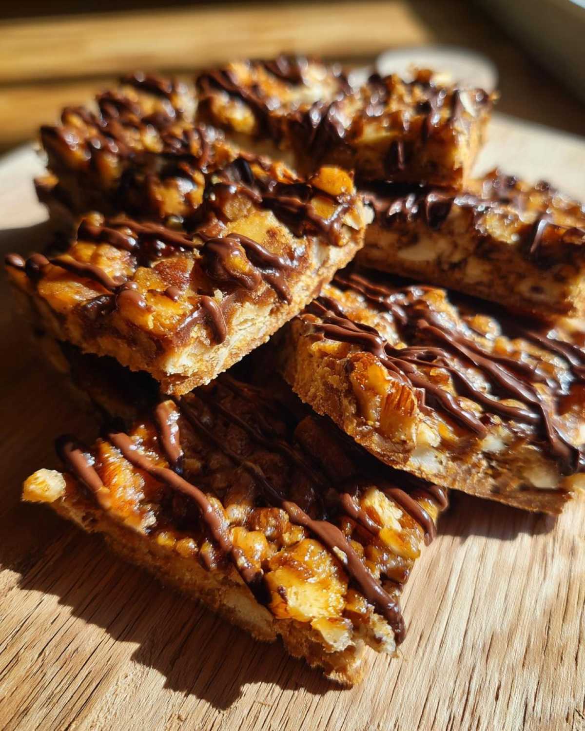 Several stacked slices of Pecan Pie Bark, featuring a nutty caramel topping drizzled with dark chocolate, sitting on a wooden surface.