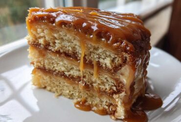 Close-up of a moist slice of caramel cake with three layers and thick, glossy caramel dripping down the sides.