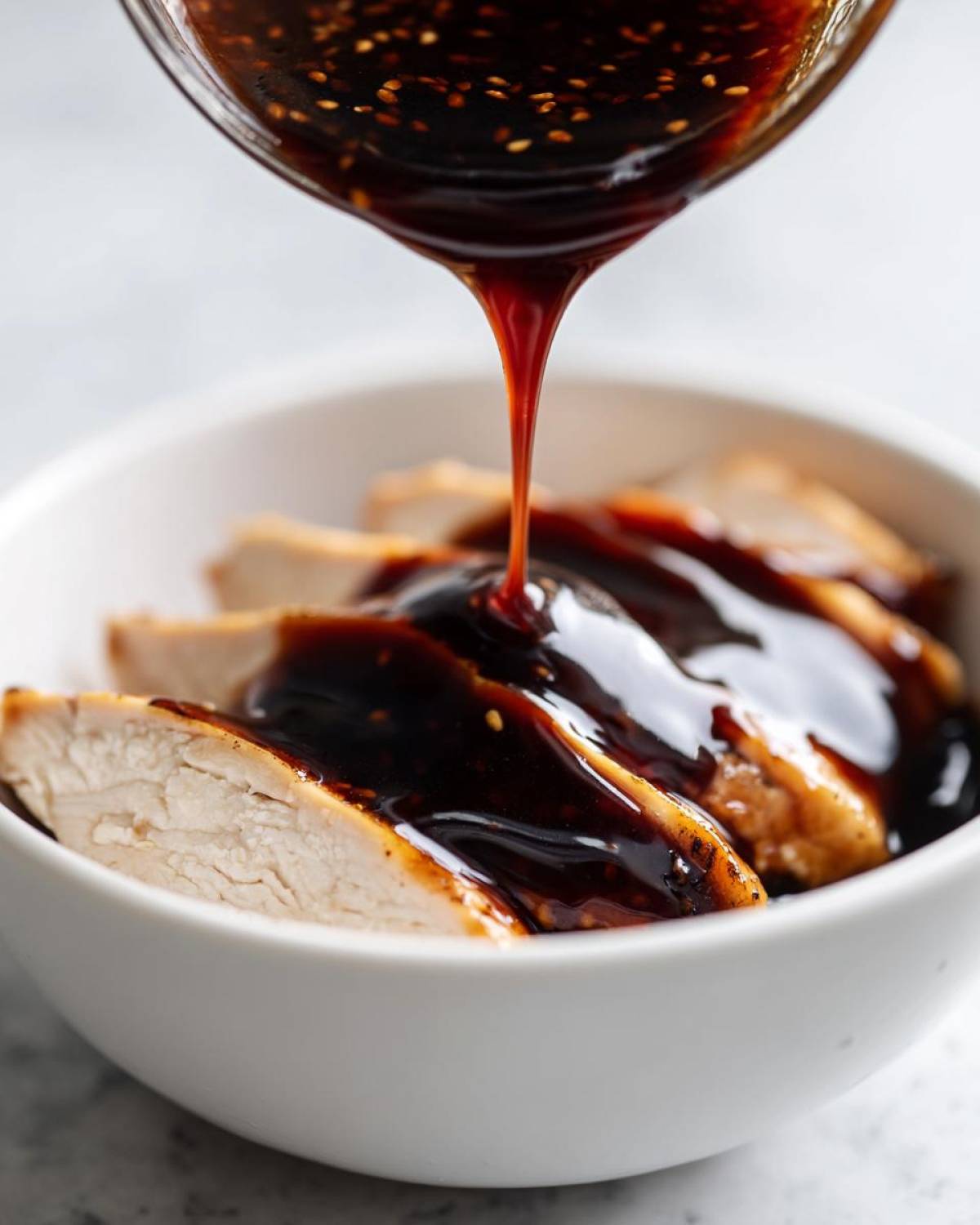 Thick, glossy homemade teriyaki sauce being poured from a glass container onto sliced, cooked chicken breast in a white bowl.