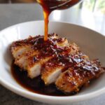 Thick, glossy teriyaki sauce being poured over slices of crispy, breaded pork cutlet in a white bowl.