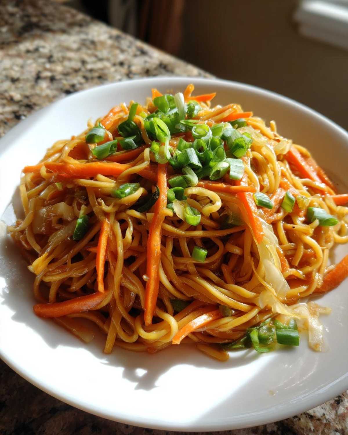 A close-up of a white bowl filled with savory fried noodles, julienned carrots, and topped with fresh green onions.