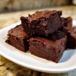 Close-up of several rich, dark chocolate fudge squares stacked on a white plate.
