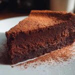 A decadent slice of flourless chocolate torte, intensely dark and dusted heavily with cocoa powder, served on a white plate.