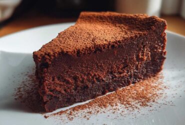 A decadent slice of flourless chocolate torte, intensely dark and dusted heavily with cocoa powder, served on a white plate.