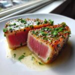 Two thick slices of perfectly seared tuna steak showing a rare, pink center, topped with herbs and sauce.