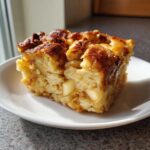 A square serving of moist apple bread pudding, topped with cinnamon sugar, on a white plate.